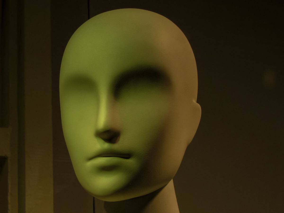 A smooth, featureless sculpted head lit in green against a dark wall.