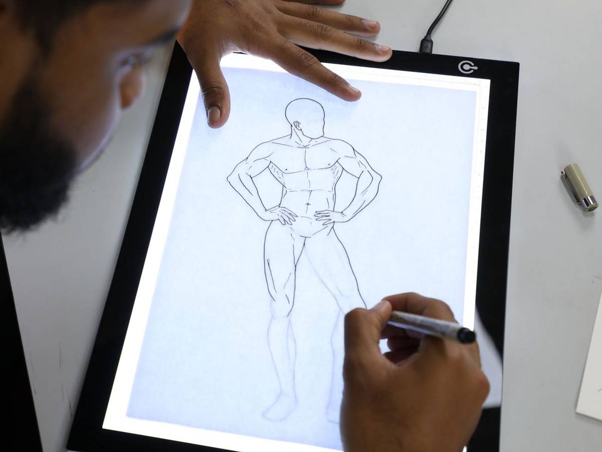 A pen tracing a standing figure drawing on a backlit light pad, one hand holding the paper steady.