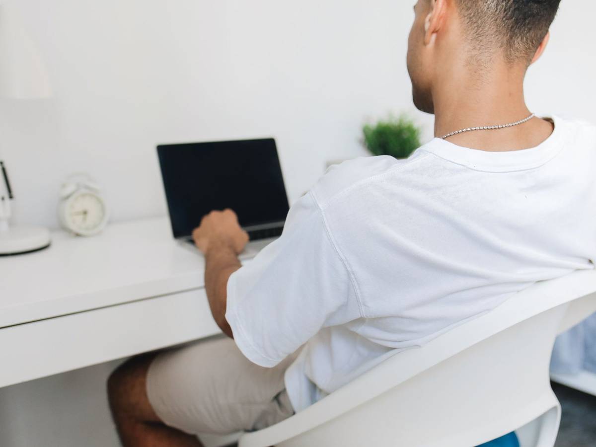 A student seen from behind working on a laptop at a small desk at home.