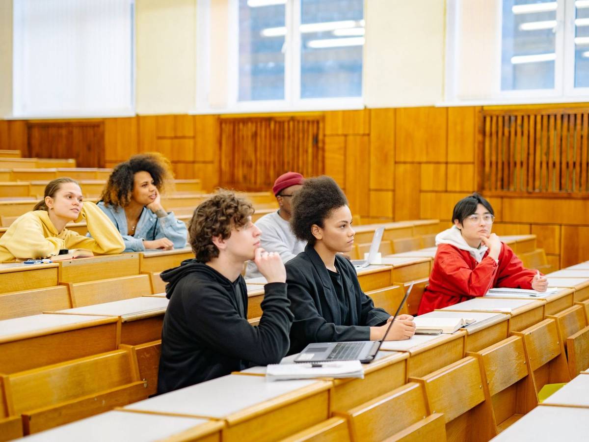 Students with laptops and notebooks sit at long wooden desks in a large tiered university lecture hall.