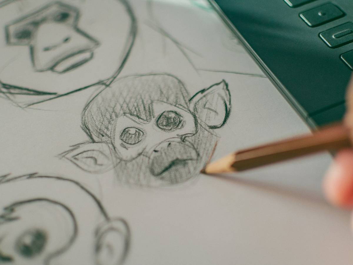 A hand holding a pencil shades a sketch of a big-eyed monkey face on paper, beside other rough character sketches and the edge of a drawing tablet.