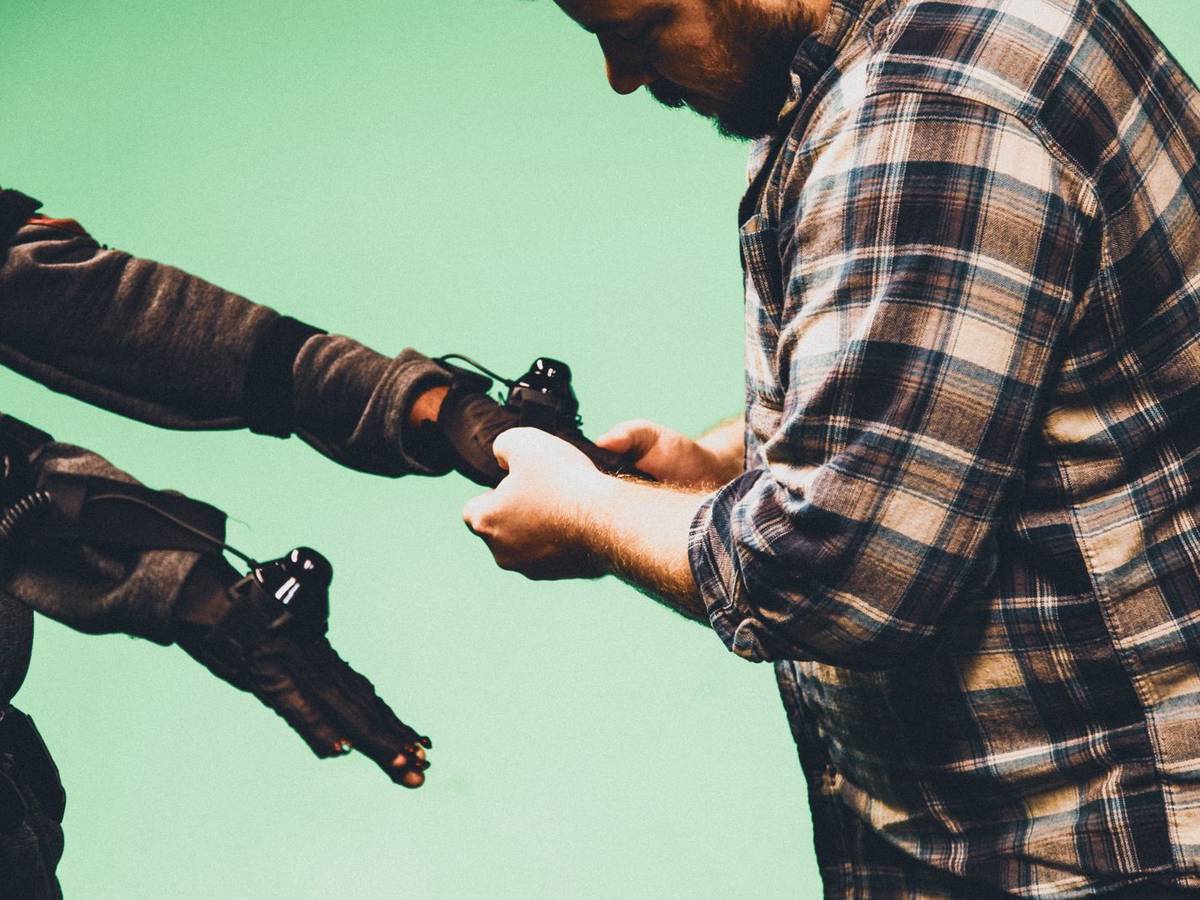 A bearded man in a plaid shirt adjusts a motion capture sensor on the gloved hand of a performer in a dark capture suit, in front of a green screen.