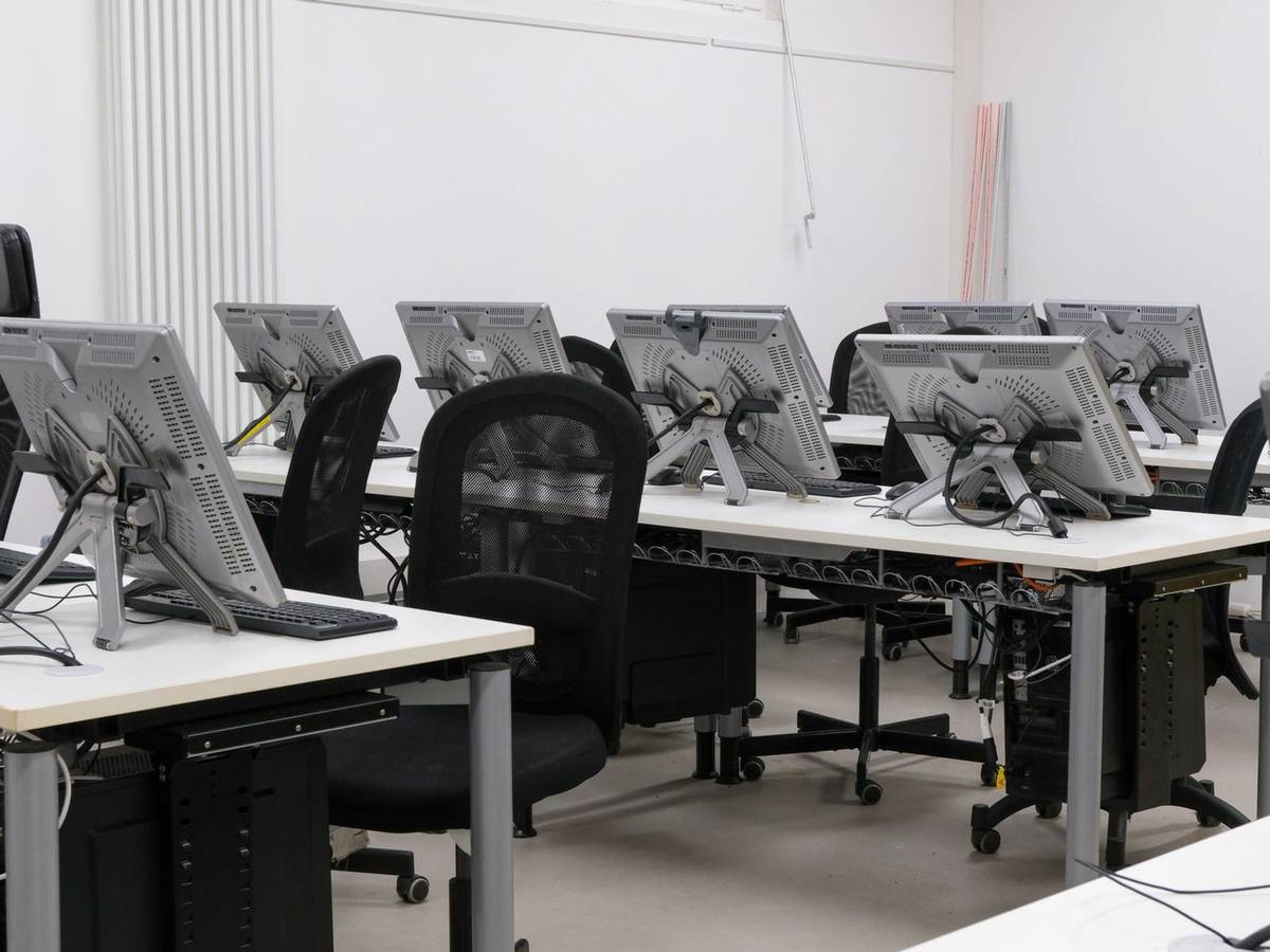 Rows of white desks in a bright classroom, each with a pen-tablet monitor and keyboard, seen from behind with empty chairs.