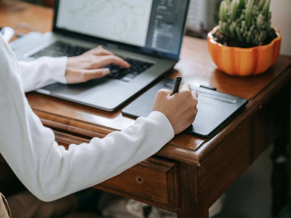 A person in a white sweater drawing with a stylus on a graphics tablet while typing on a laptop at a wooden desk.