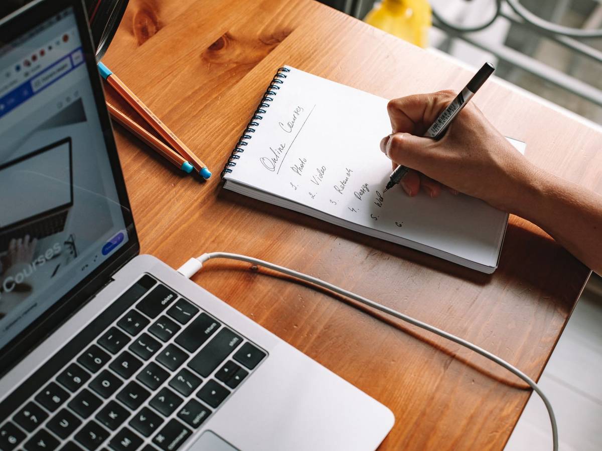 A hand writing a numbered list headed Online Courses in a spiral notebook next to an open laptop on a wooden desk.