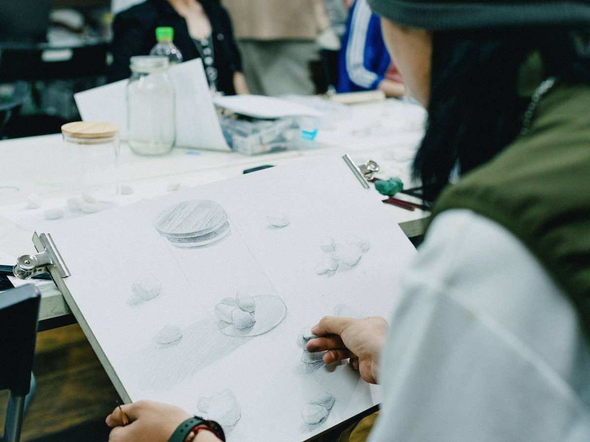 An art student in a studio classroom shades a pencil study of glass objects on a sheet clipped to a drawing board, with classmates at work behind.