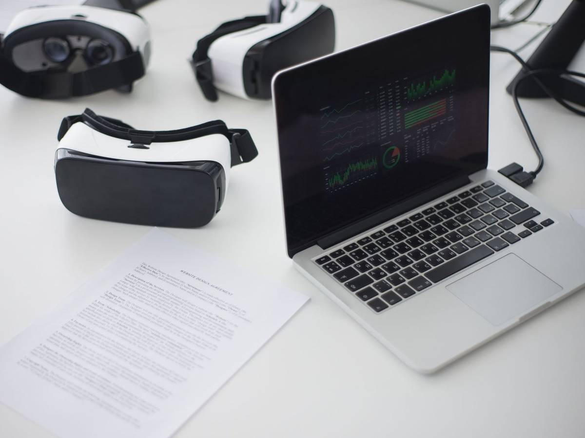 Three virtual reality headsets on a white desk beside an open laptop and printed notes.