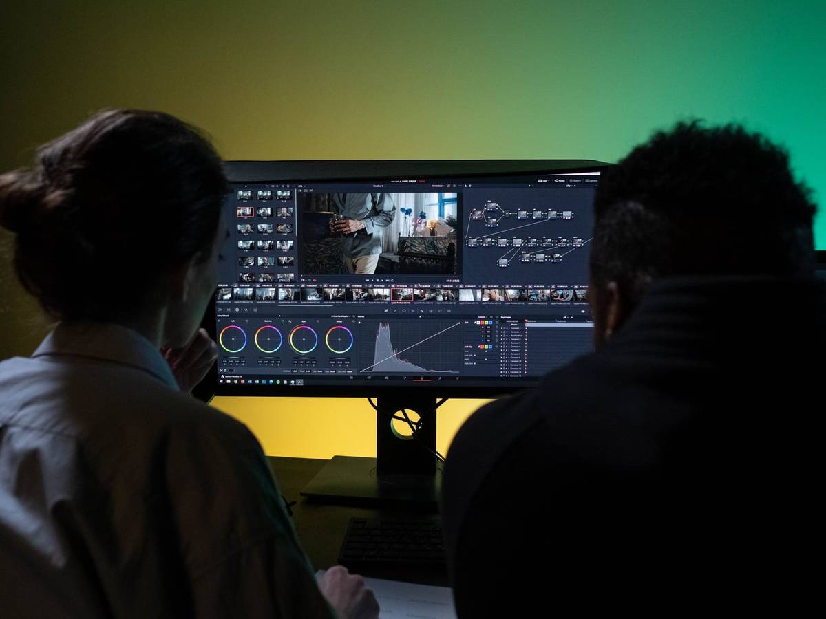 Two people seen from behind reviewing a video edit on a wide monitor, its timeline of clips and color wheels lit against a green and yellow wall.