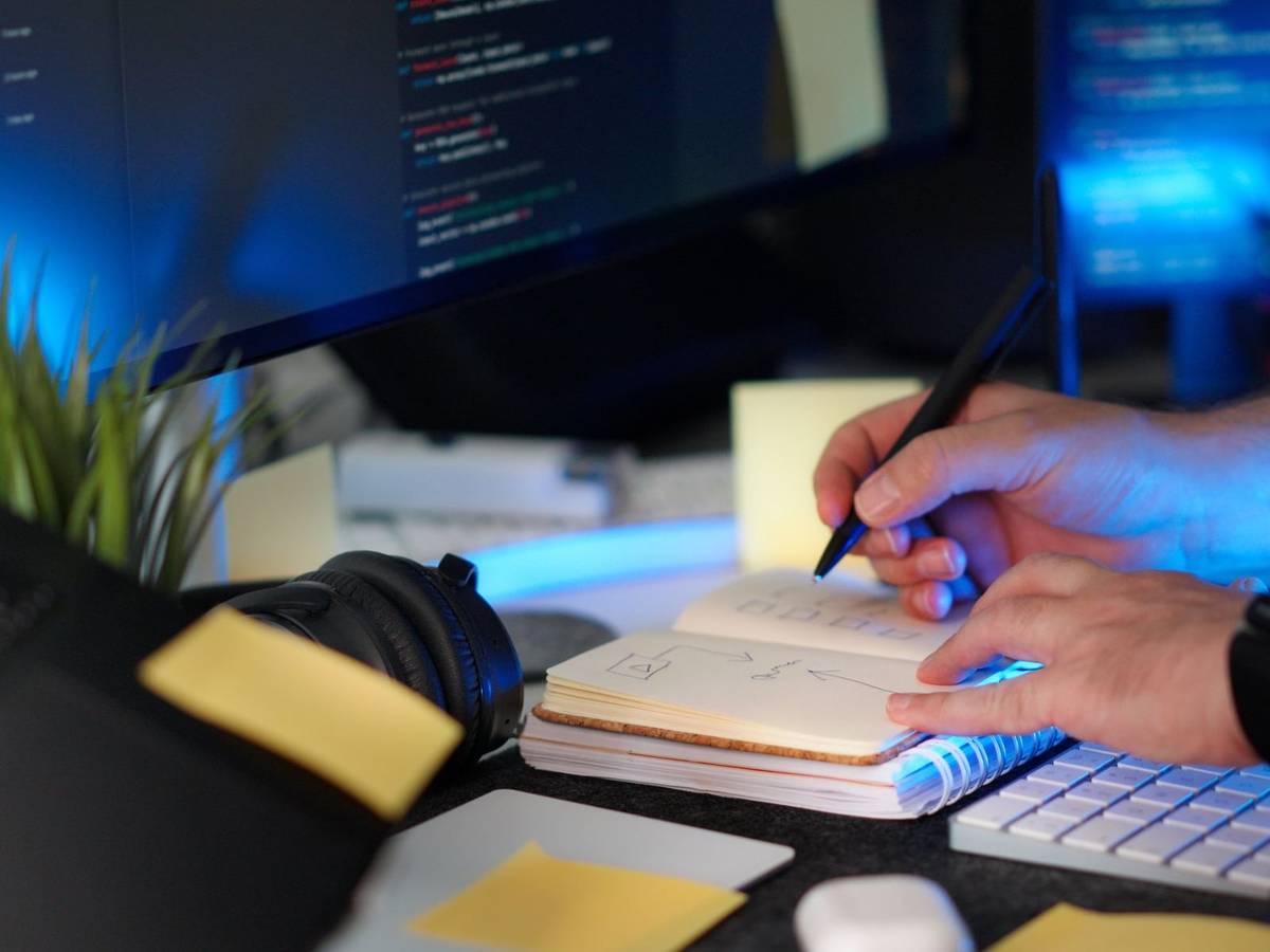 A hand sketching in a small notebook beside a keyboard, with lines of code glowing on a blue-lit monitor behind it.