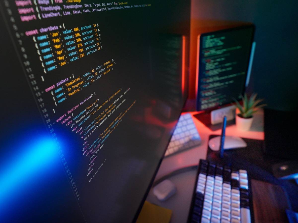 Colorful source code in a code editor across two monitors at a programmer's desk lit in blue and red.