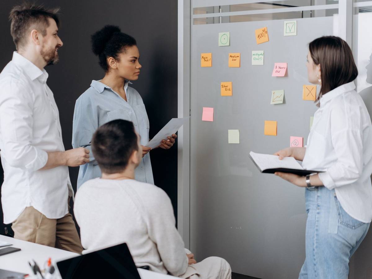 Four colleagues gathered at a gray wall covered in sticky notes, one holding a notebook, during a planning meeting.
