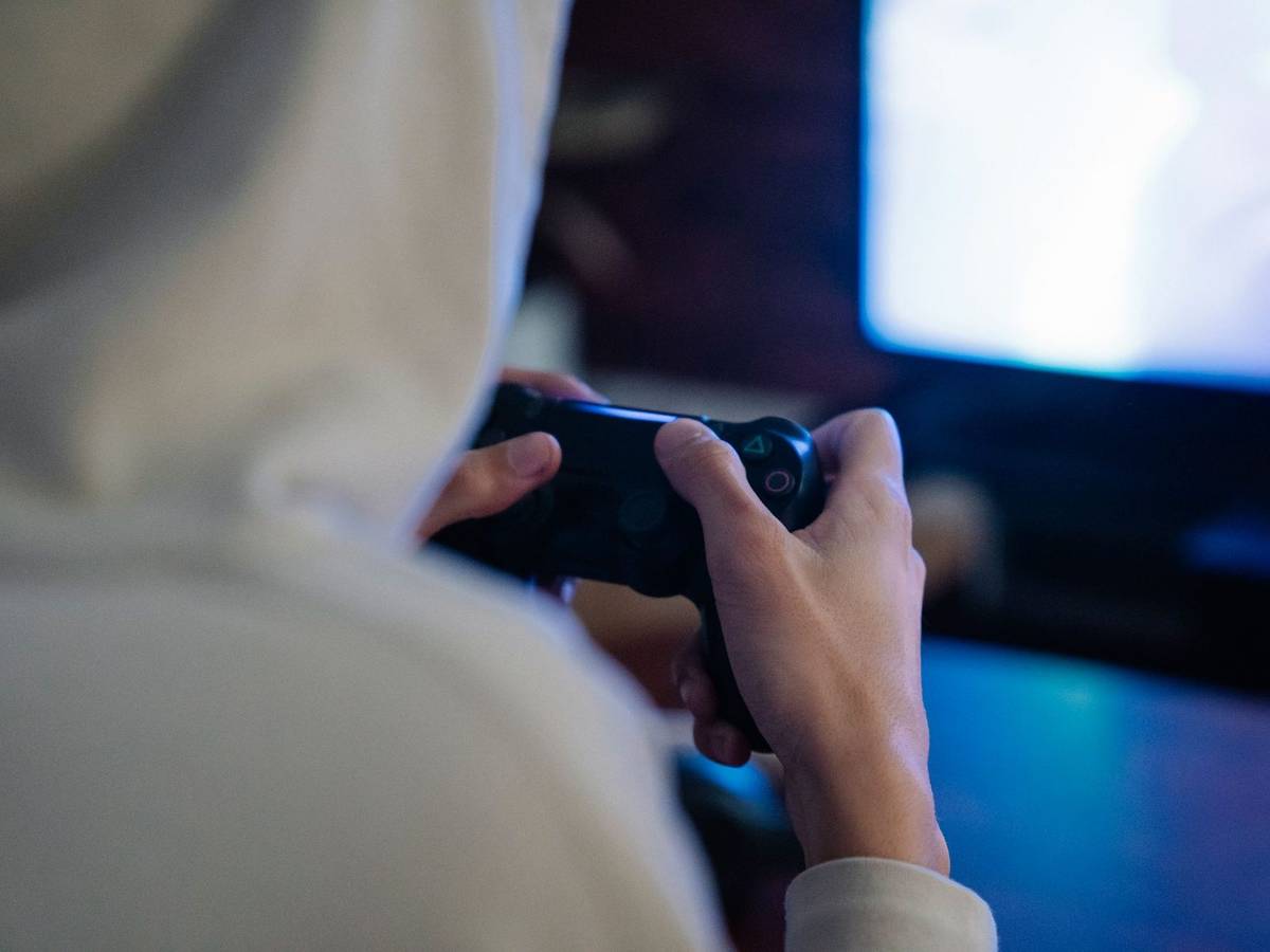 A person seen from behind in a hooded top holding a game controller in front of a glowing screen in a dim room.