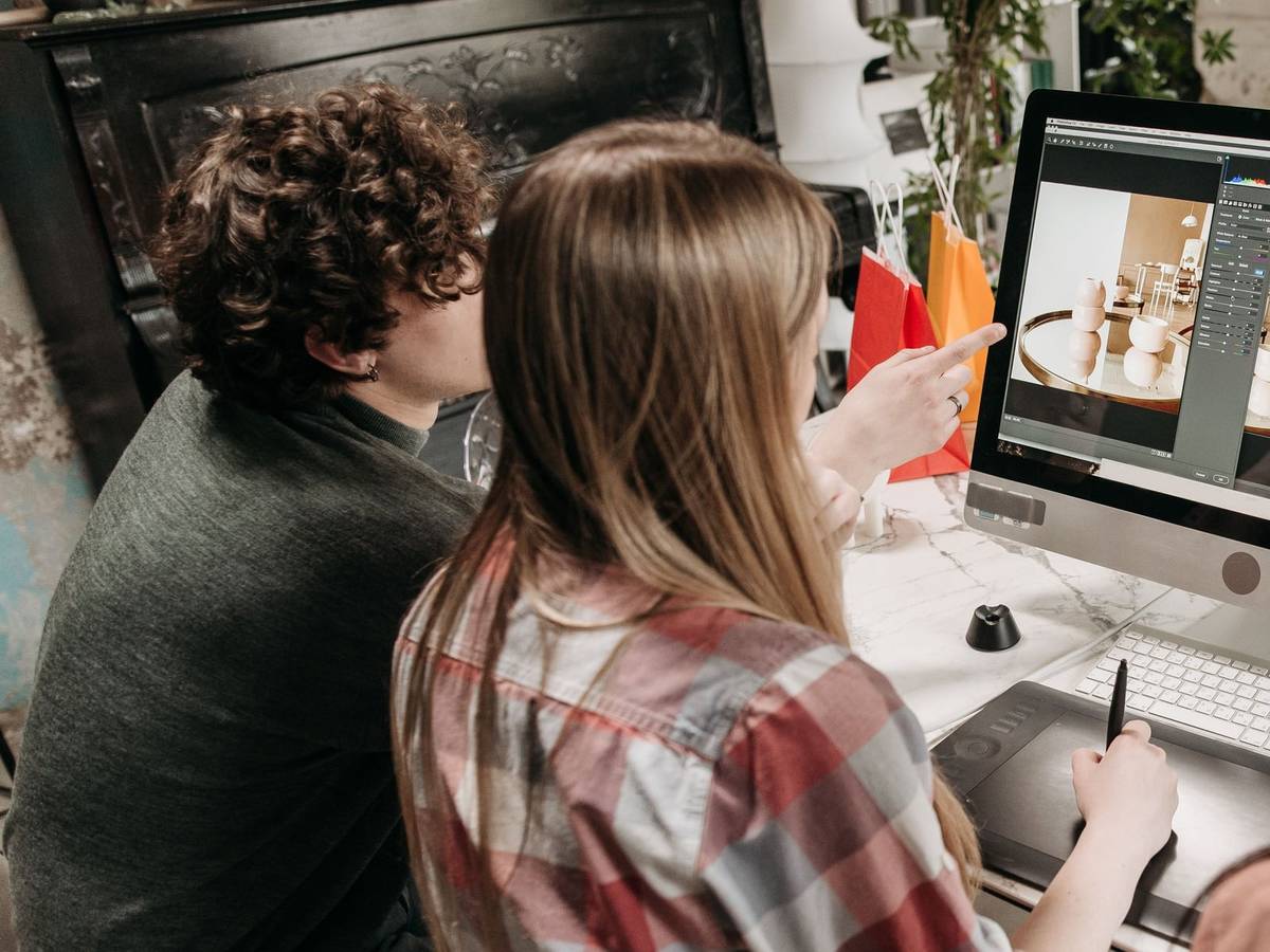 Two colleagues seen from behind at a desktop computer, one pointing at an image on screen while the other works on a drawing tablet.