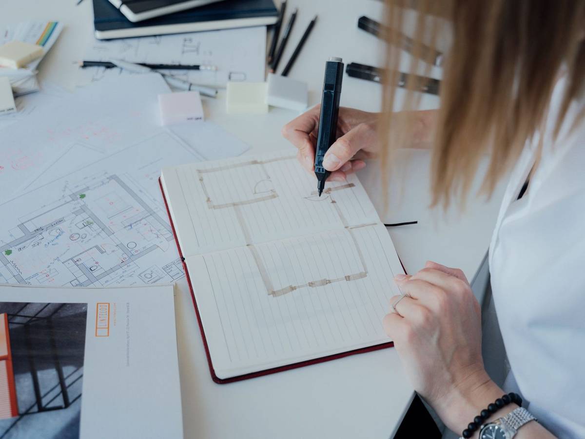 A hand sketching a floor plan with a marker in a notebook, surrounded by printed layout drawings.