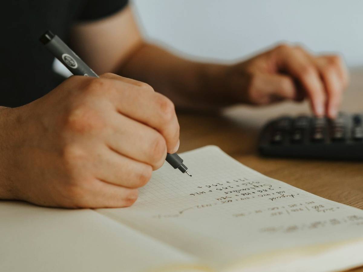 Close-up of a hand writing monthly sums in a grid notebook while the other hand uses a calculator on a wooden desk.