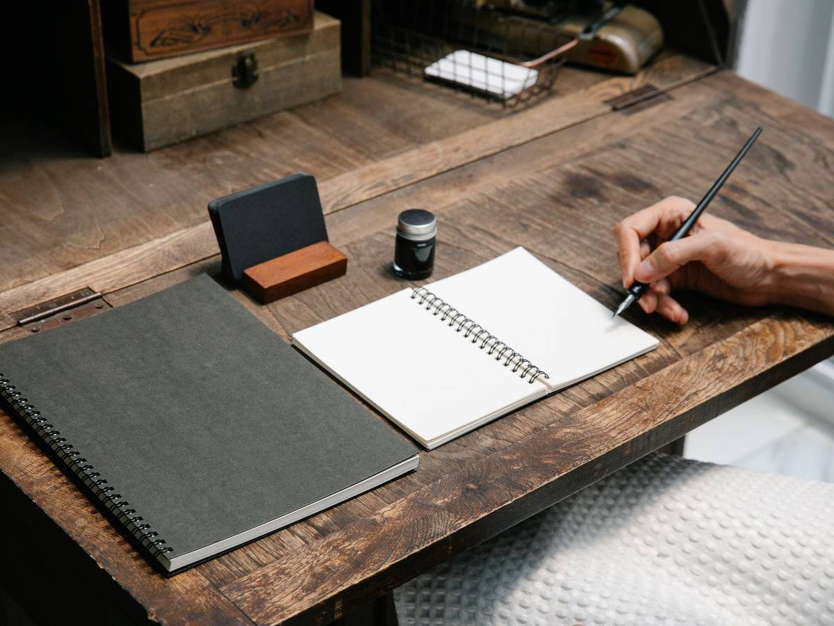 A hand writing with a pencil in an open spiral notebook on a rustic wooden desk beside a closed notebook and an inkwell.