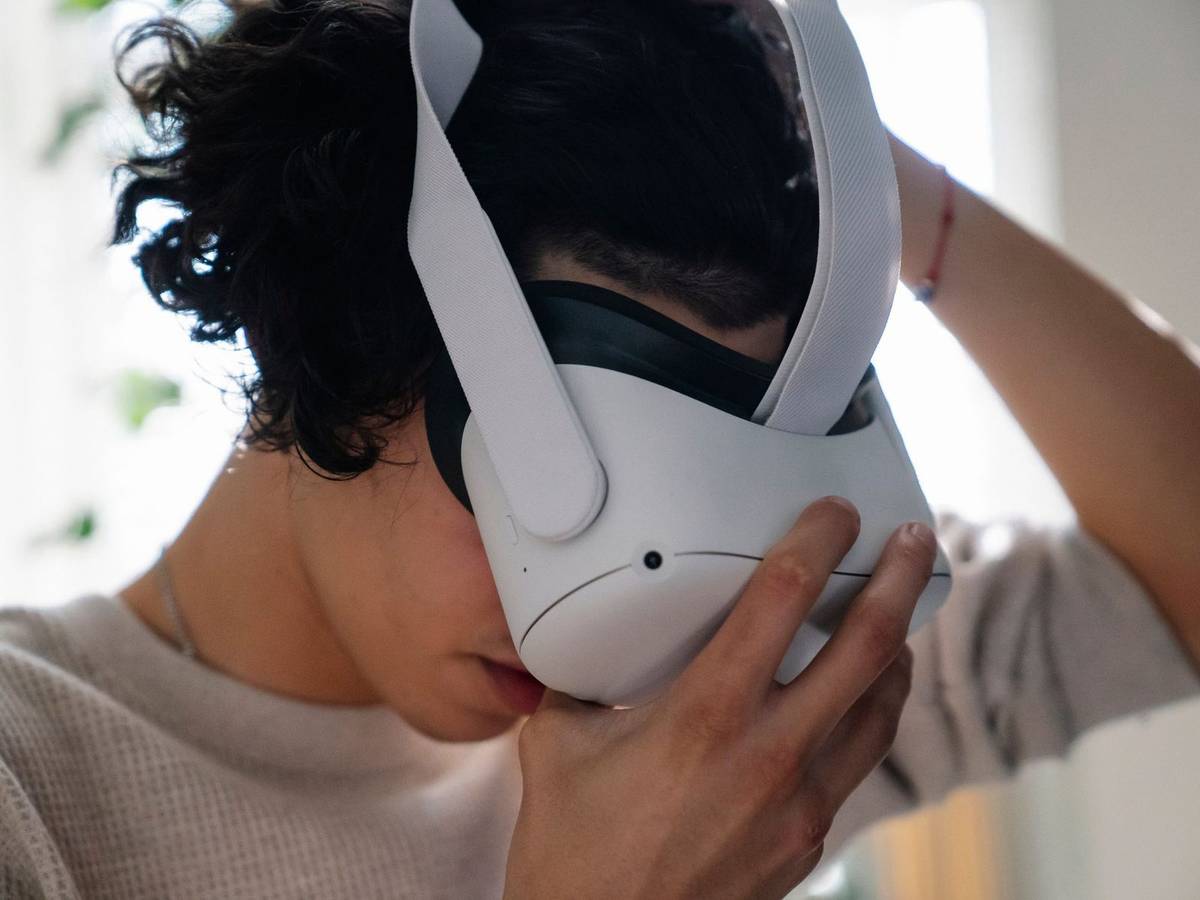 A person in a light sweater lifting a white virtual reality headset onto their face, by a bright window.