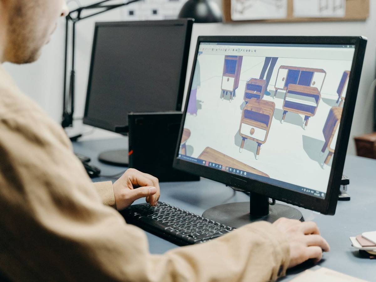 A person at a desktop workstation arranging a 3D model of cabinets in modeling software on the monitor.