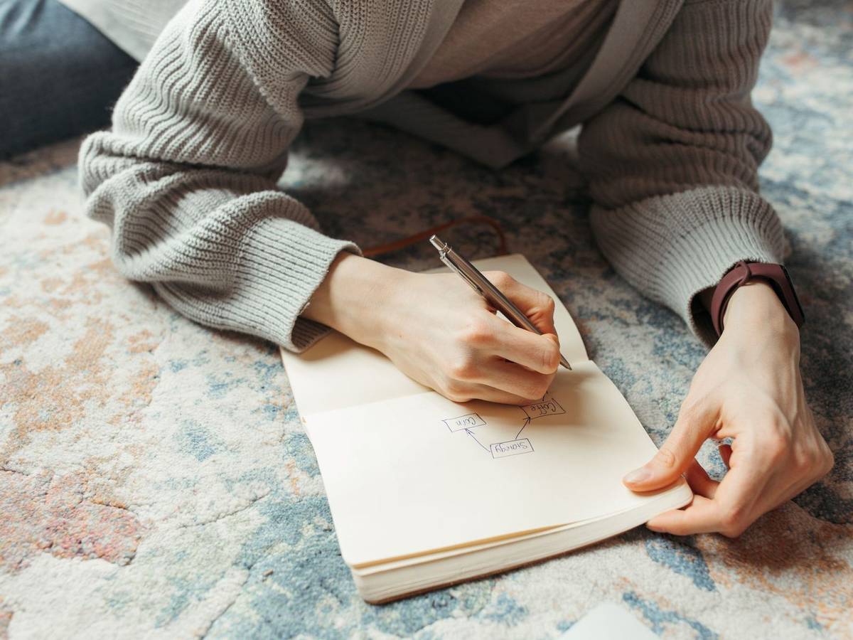 A person lying on a rug drawing a small flowchart in an open notebook with a pencil.