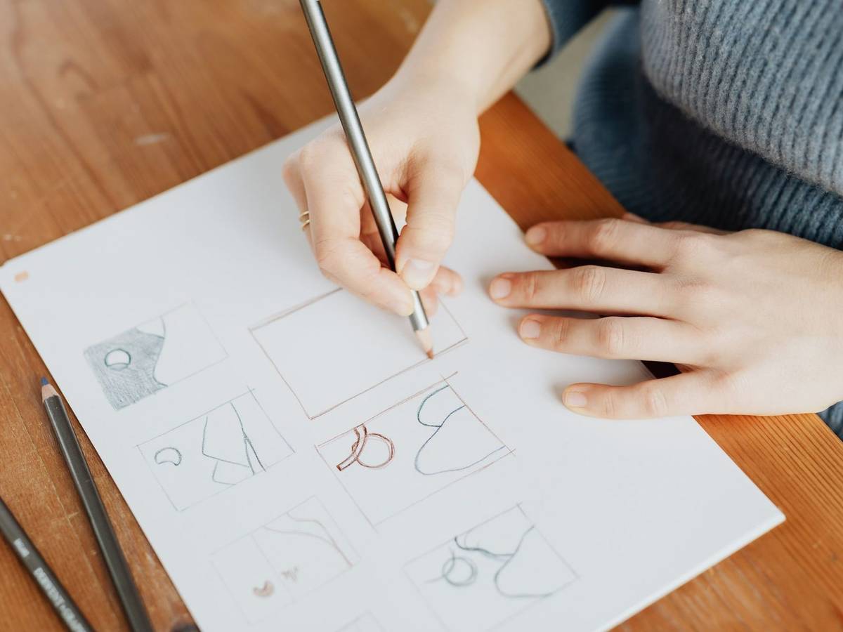 A hand drawing a grid of small frame sketches in pencil on a sheet of paper at a wooden desk.