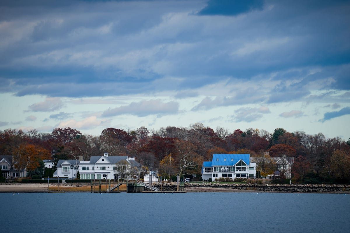 Large waterfront houses behind trees in autumn color, seen across calm water under a soft sky