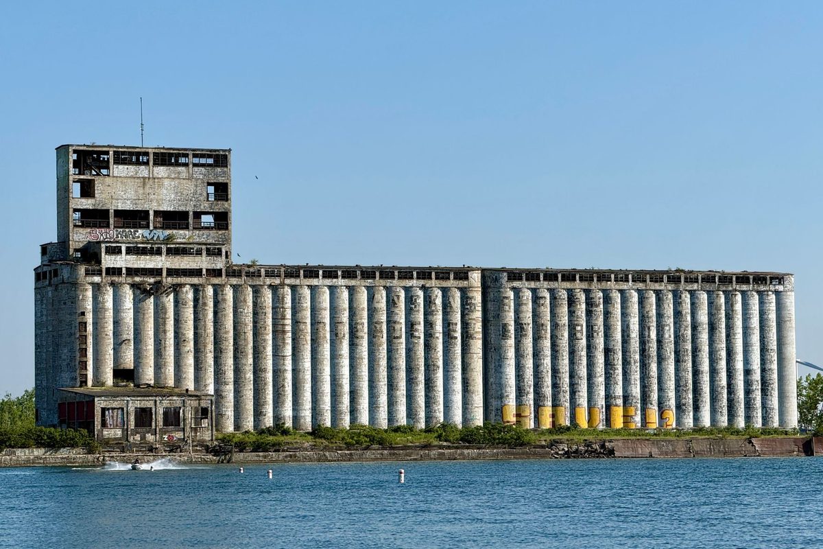 A long row of old concrete grain silos with a tall elevator tower on a waterfront, beside blue water under a clear sky