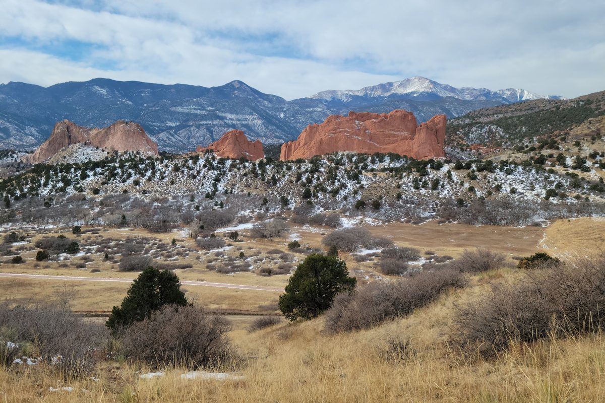 Jagged red sandstone rock formations rise from dry golden grassland dotted with juniper, with a snow-capped peak and forested ridges behind