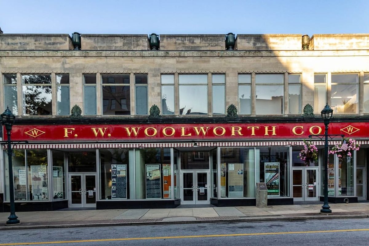 A two-story pale stone commercial building on a quiet downtown street, with a long red storefront sign, tall windows and black lampposts under a clear sky