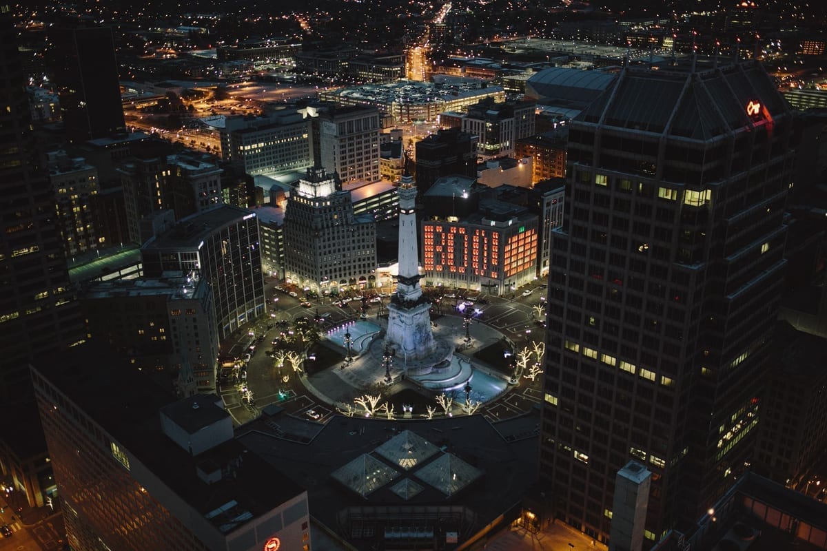 A circular plaza with a lit monument at night, seen from above and ringed by towers