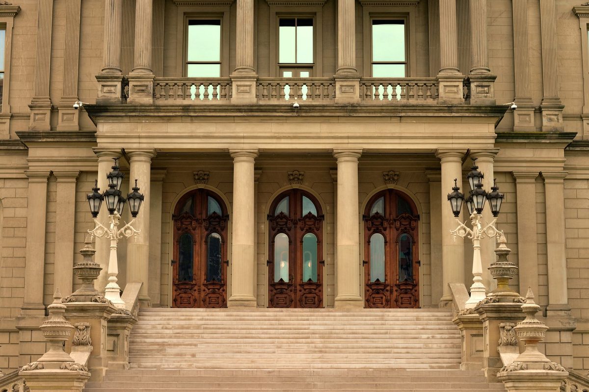 A columned stone entrance with three tall arched wooden doors, a balcony above and ornate lamp posts either side of wide steps