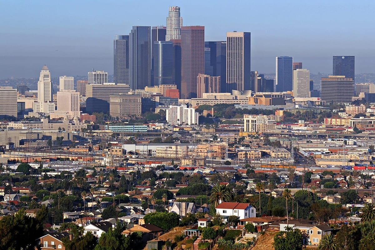 A cluster of downtown towers in haze beyond a hillside of low houses, palm trees and rooftops