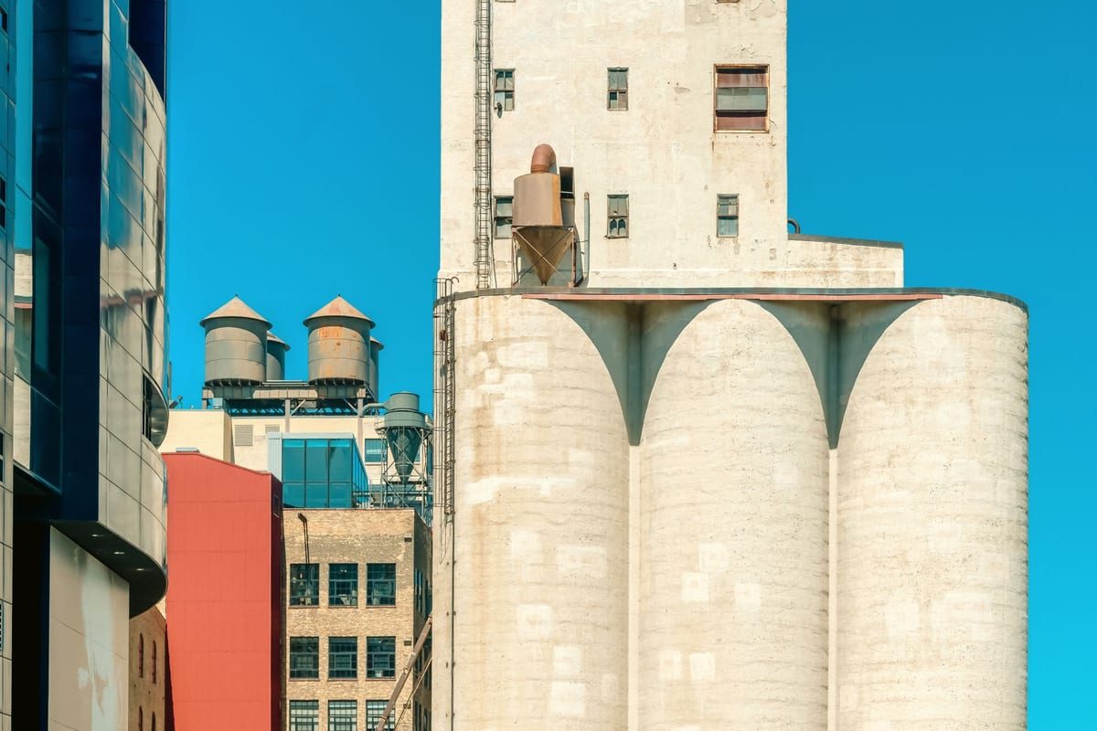 Concrete grain silos and a mill tower in pale stone beside a red brick warehouse and blue glass, under a hard blue sky