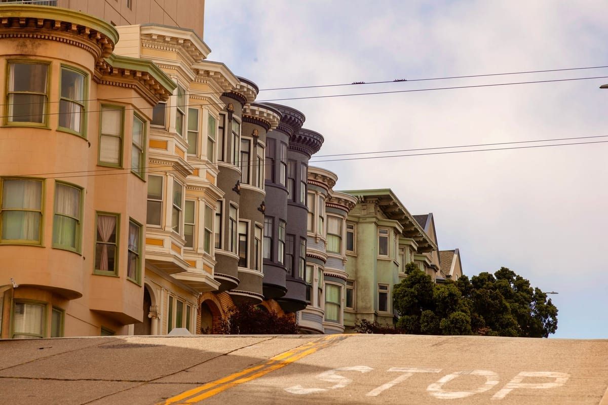 A row of bay-windowed townhouses on a steep street, with a stop marking painted on the road