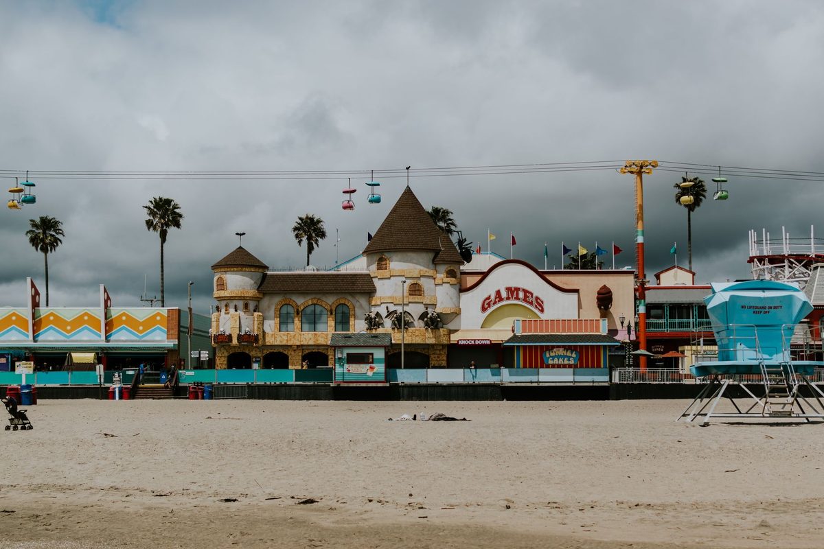 A castle-style arcade and colorful boardwalk buildings behind a sandy beach and a blue lifeguard tower, under heavy gray clouds