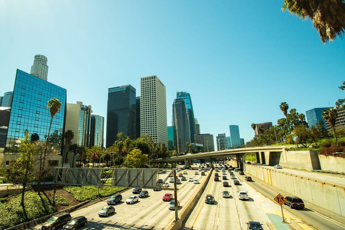 A freeway full of traffic running between glass office towers and palm trees under a clear sky