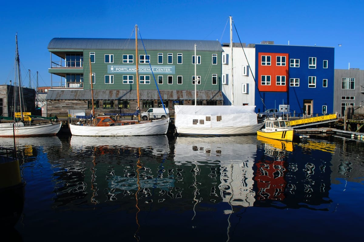 Wooden sailboats tied up at a pier in front of green and blue harbor buildings