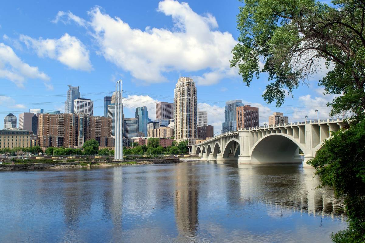 A city skyline across a river, a stone arch bridge and green trees at its edge