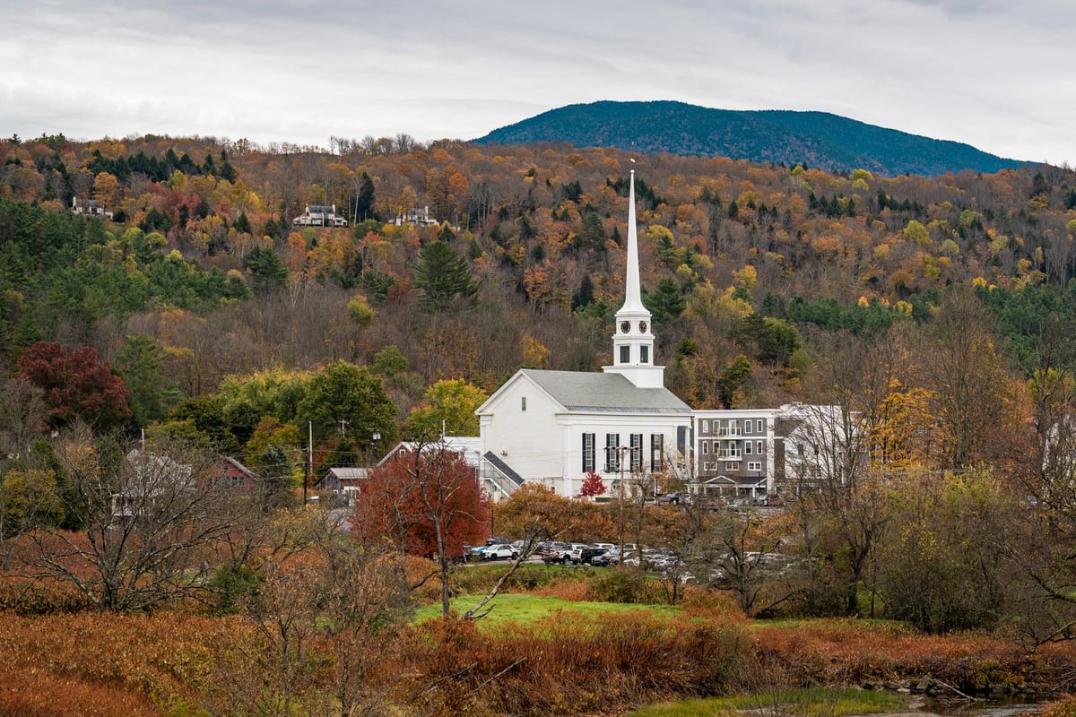 A white church steeple among autumn trees below a rounded green mountain