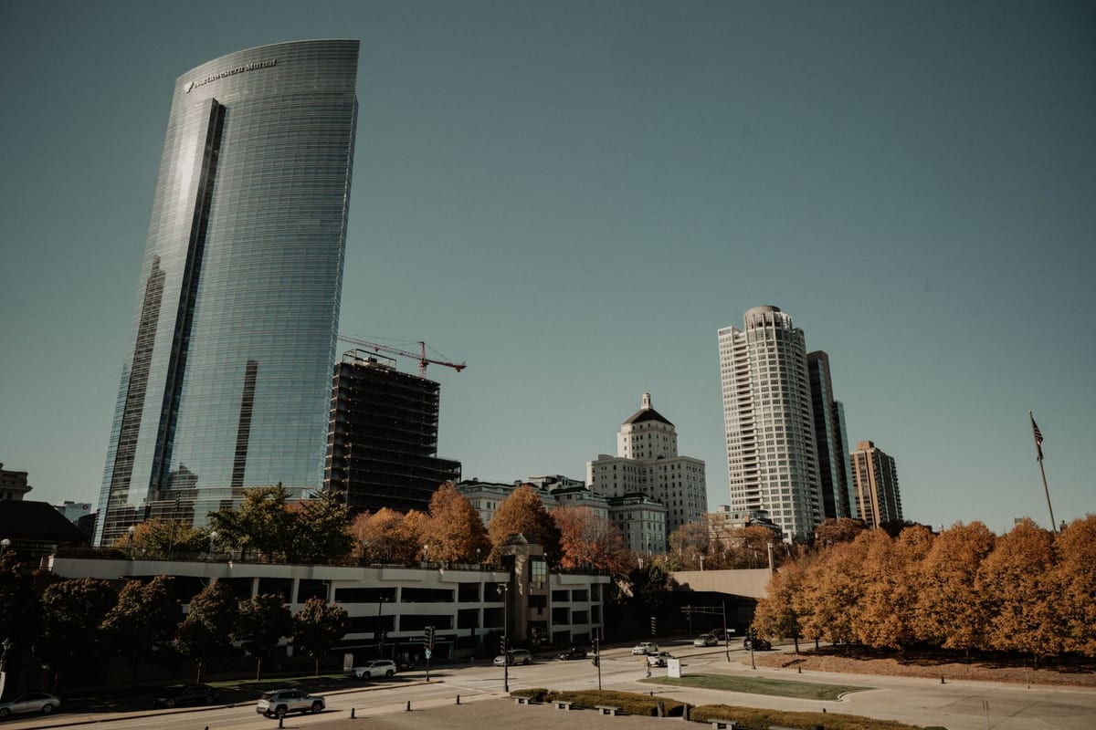 A tall curved glass tower beside smaller buildings, autumn trees along a lawn